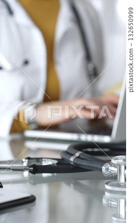 Stethoscope and clipboard with medical documents are on the table near female doctor working on laptop, managing patient data in clinic. Concept of medicine, healthcare and science 132650999