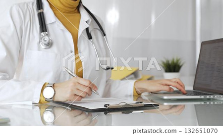 Doctor wearing stethoscope and yellow sweater under white lab coat, making notes on a clipboard in a bright medical office. Medicine, healthcare and science concept 132651070