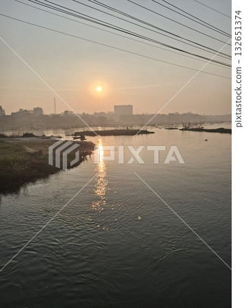 Serene sunrise over a calm river, with reflections shimmering on the water and a distant cityscape 132651374