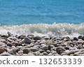 Smooth, grey, and white pebbles fill the foreground with a sparkling blue sea and a breaking wave in the blurred background. Concept of serene beach scenery and natural textures. 132653339