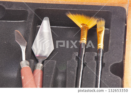 Close-up of a set of art tools: two metal palette knives with red handles and two brushes with gold ferrules and black handles. The tools are neatly arranged in a black wooden case. 132653350
