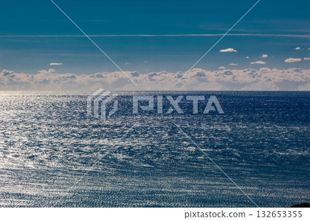 A wide seascape with a clear horizon line. The deep blue surface of the water reflects bright sun glare. A horizontal strip of white clouds stretches across the horizon.  132653355