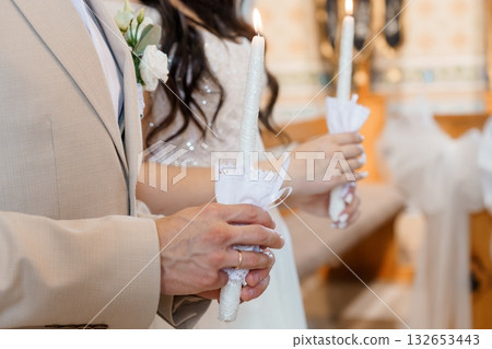 Wedding couple holding lit unity candles during ceremony in church with floral boutonniere and lace accents Wedding couple holding lit unity candles during ceremony in church with floral boutonniere and lace accents 132653443