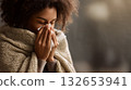 Person Wrapped in Beige Knitted Blanket Holding Tissue to Nose Indoors with Warm Lighting 132653941