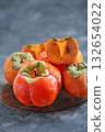 A platter holds several vibrant orange persimmons, one of which is cut in half revealing its juicy interior, set against a textured grey background. Concept of fresh, healthy fruit. 132654022