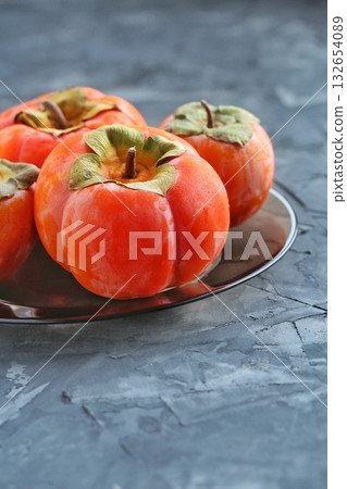 A group of vibrant orange persimmons, some with visible water droplets, are artfully arranged on a dark plate against a textured grey background. Concept of fresh, healthy autumn fruit. A group of vibrant orange persimmons, some with visible water droplets, are artfully arranged on a dark plate against a textured grey background. Concept of fresh, healthy autumn fruit. 132654089