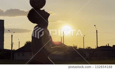 Young man juggling soccer ball on stadium at sunset. Professional footballer kicking ball at green field. Sportsman practicing tricks at meadow with sunlight at background. Freestyle football Young man juggling soccer ball on stadium at sunset. Professional footballer kicking ball at green field. Sportsman practicing tricks at meadow with sunlight at background. Freestyle football 132654188