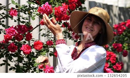 Portrait of adult smiling woman wearing a traditional ukrainian vyshyvanka among blooming red roses in summer garden. This scene expresses femininity, cultural heritage and national identity 132654205