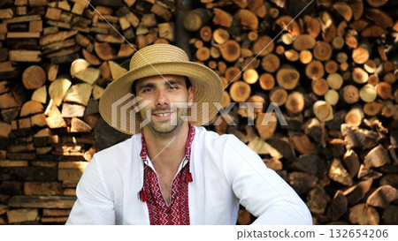 Portrait of ukrainian man in straw hat against the background of woodshed. Young smiling guy wearing an embroidered shirt looking into camera outdoor. Concept of national identity and independence 132654206