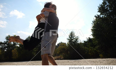 Happy couple having fun together on seashore at summer vacation. Man taking woman in air and turning around at coastline. Carefree lovers spending time at sandy beach. Concept of love. Slow motion 132654212