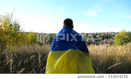 Military man in uniform stands with flag of Ukraine on shoulders at countryside. Portrait of male ukrainian soldier with national banner in honor of victory against russian aggression. End of war 132654226