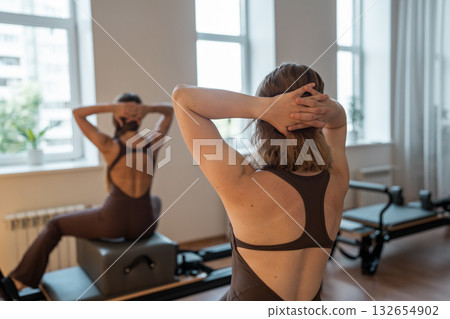 Fit women in black sportswear doing pilates side plank exercises on pilates reformer machine bed in minimal bright modern studio.  132654902