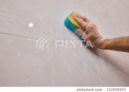 A Man Diligently Cleaning a Tiled Bathroom Wall with His Gloves and a Scrubber Tool 132656843