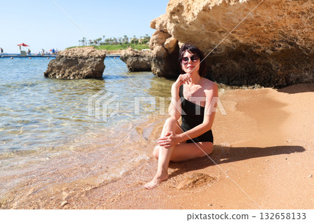 A brunette woman with glasses in a black bathing suit sits on the sand overlooking the sea, with her legs tucked under her, enjoying the sun's rays 132658133