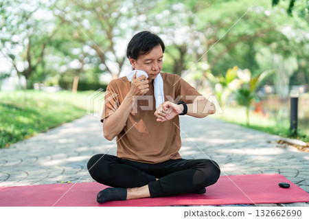 Mindful Fitness. Senior man checking watch while resting on yoga mat in a serene park. 132662690