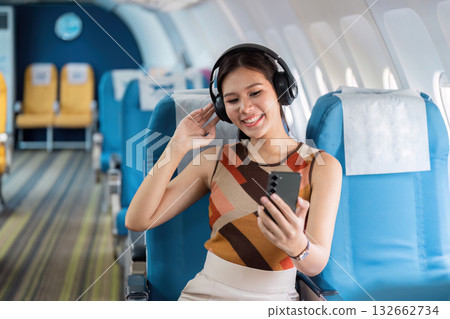 Tourist and Inflight Entertainment. A woman with headphones smiling while using her smartphone on an airplane. Tourist and Inflight Entertainment. A woman with headphones smiling while using her smartphone on an airplane. 132662734