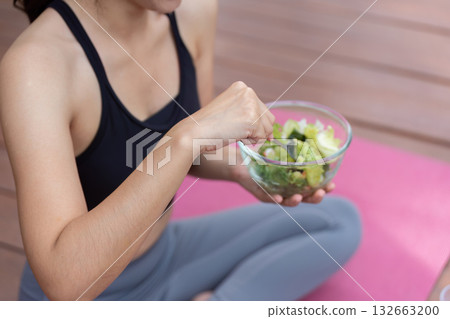 Healthy Eating and Wellness. Woman enjoying fresh salad while seated on exercise mat, promoting nutrition. Healthy Eating and Wellness. Woman enjoying fresh salad while seated on exercise mat, promoting nutrition. 132663200