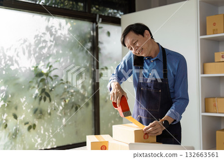 E-commerce professional happily packing boxes with tape by a large window. E-commerce professional happily packing boxes with tape by a large window. 132663561