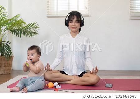 Mindful mother meditating with headphones while her baby plays nearby, promoting wellness and tranquility in a serene indoor space. 132663563