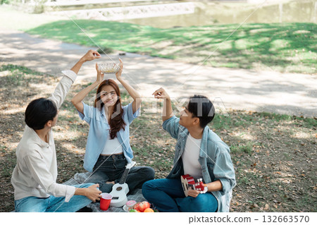 Friends playfully sharing snacks at a picnic while enjoying music in a park. 132663570