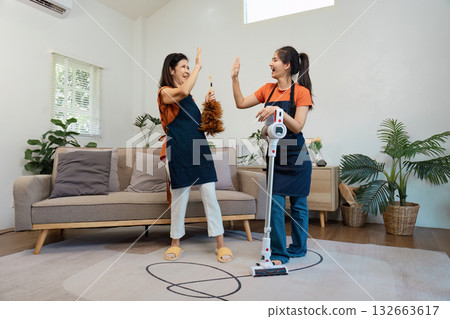 Two women giving a high-five while cleaning a living room, one with a feather duster and the other with a vacuum cleaner. 132663617