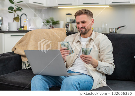 Young man counts cash dollars compares with laptop earnings sheet fills sums and smiles at goal met 132666350