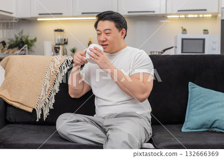 Asian man holds cup of herbal tea warms hands smiles breathes aroma enjoys pause mindful calm break Asian man holds cup of herbal tea warms hands smiles breathes aroma enjoys pause mindful calm break 132666369