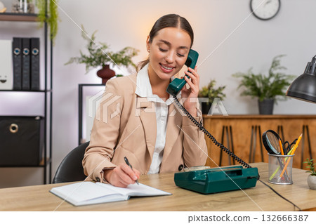 Young businesswoman answers retro landline from nineties speaks clear business talk and writes notes Young businesswoman answers retro landline from nineties speaks clear business talk and writes notes 132666387