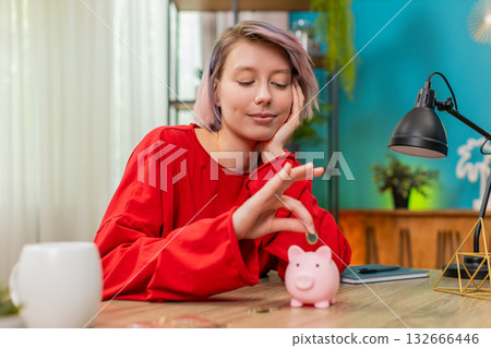 Young woman happily putting coins into piggy bank smiling expressing financial discipline budgeting 132666446