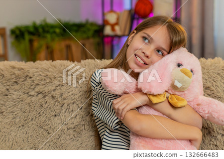Happy child girl kid hugging favorite bunny toy gently with smile while sitting on cozy home sofa 132666483