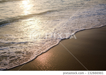 Peaceful ocean waves gently wash over golden sand at sunset, creating a serene and tranquil beach atmosphere with shimmering reflections 132666981