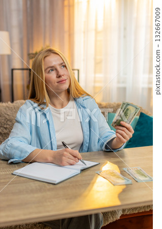 Young woman organizing expenses at home, counting money cash banknotes, writing notes, happy focus 132667009