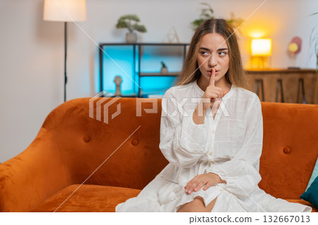 Portrait of young woman on sofa presses index finger to lips makes silence hush gesture sign at home Portrait of young woman on sofa presses index finger to lips makes silence hush gesture sign at home 132667013