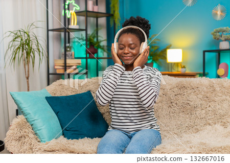 African American woman girl enjoying listening music, dancing on sofa, wireless headphones at home African American woman girl enjoying listening music, dancing on sofa, wireless headphones at home 132667016