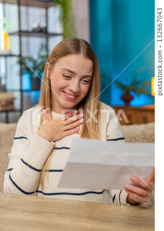 Caucasian young woman receiving important letter at home on sofa, opening envelope, feeling joyful 132667043
