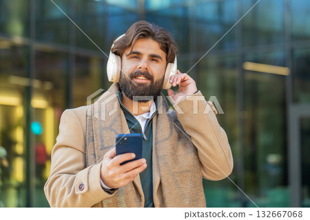 Happy young man putting on headphones listening favorite energetic music on smartphone on street Happy young man putting on headphones listening favorite energetic music on smartphone on street 132667068