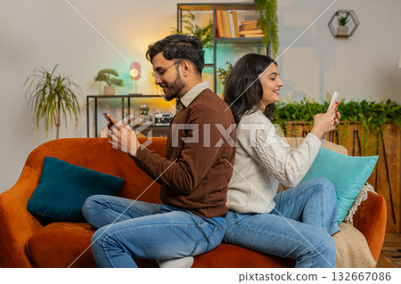 Side view of cheerful diverse couple sitting back to back using smartphones talking on home sofa Side view of cheerful diverse couple sitting back to back using smartphones talking on home sofa 132667086