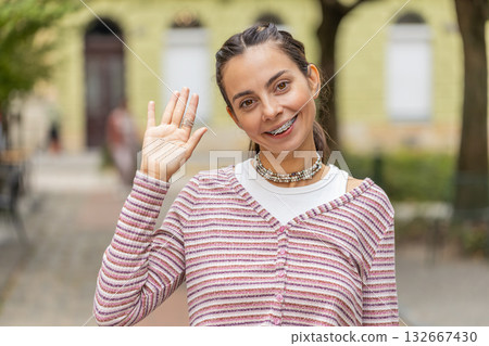 Happy woman smiling friendly at camera waving hands gesturing hello greeting goodbye on city street Happy woman smiling friendly at camera waving hands gesturing hello greeting goodbye on city street 132667430