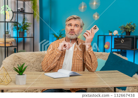 Mature man at sofa table on mobile phone speaker call, writing notes in notebook, serious at home 132667438