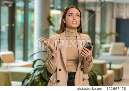 Young businesswoman using smartphone becomes shocked sudden lottery results in office hotel lobby Young businesswoman using smartphone becomes shocked sudden lottery results in office hotel lobby 132667458