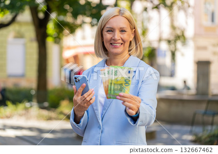 Caucasian mature businesswoman on city street showing earnings cash money with smartphone in hands Caucasian mature businesswoman on city street showing earnings cash money with smartphone in hands 132667462