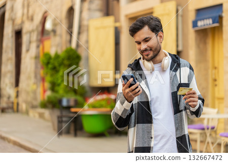 Smiling young man using credit card smartphone while transferring money purchases online outdoors Smiling young man using credit card smartphone while transferring money purchases online outdoors 132667472