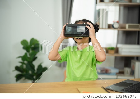 Child Engaged in Virtual Reality Experience with VR Headset while Sitting at Table in Modern Room with Laptop and Indoor Plant in Background Child Engaged in Virtual Reality Experience with VR Headset while Sitting at Table in Modern Room with Laptop and Indoor Plant in Background 132668073