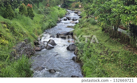 A rapid river with strong currents flows over and around large rocks, creating white water and a small cascade, surrounded by a green. A rapid river with strong currents flows over and around large rocks, creating white water and a small cascade, surrounded by a green. 132669721