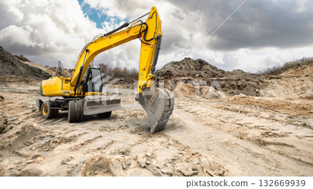 A bright yellow excavator works diligently on a rugged construction site, shaping the earth beneath a moody sky filled with swirling clouds 132669939