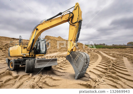 A yellow excavator maneuvers through a construction site, skillfully reshaping the sandy terrain as overcast skies loom above on a quiet afternoon 132669943