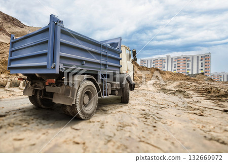 Dust swirls around a heavy truck parked on a sandy road, with modern apartments rising in the background under an overcast sky, indicating ongoing development Dust swirls around a heavy truck parked on a sandy road, with modern apartments rising in the background under an overcast sky, indicating ongoing development 132669972