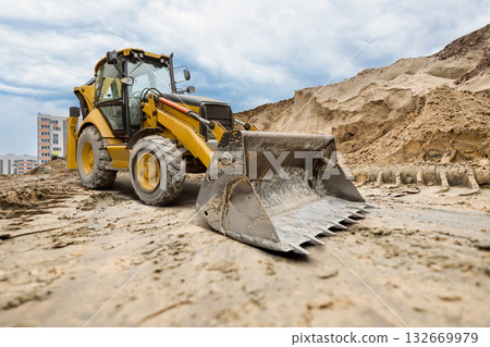 A yellow excavator moves through a construction site, skillfully digging and moving dirt against a backdrop of tall buildings and an overcast sky A yellow excavator moves through a construction site, skillfully digging and moving dirt against a backdrop of tall buildings and an overcast sky 132669979