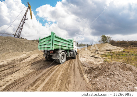 A sturdy green truck moves along a rugged dirt path beside a construction area, surrounded by mounds of earth and a crane in the distance on a cloudy day 132669983