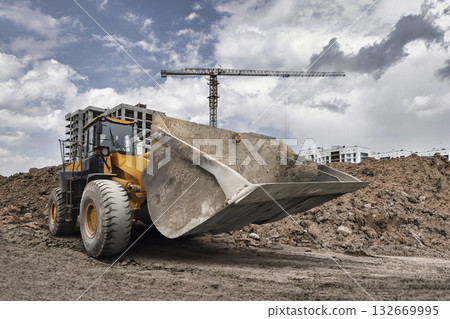 A powerful loader digs into the ground at a construction site, preparing for new buildings under a dramatic sky filled with clouds 132669995